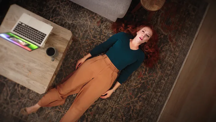 A woman lying on a living-room rug beside a laptop, laughing