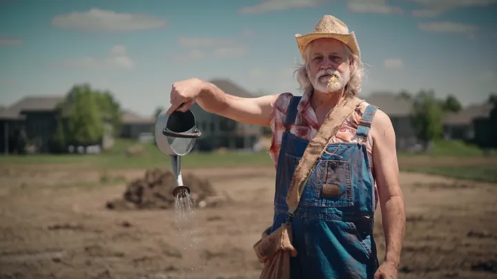 A farmer in overalls watering a bare dirt lot with a watering can, new houses behind him