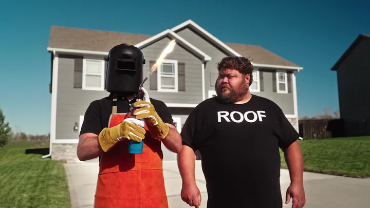 Two men outside a new house, one in a welding mask and apron