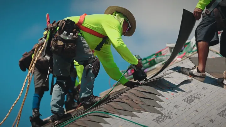 Roofers in safety harnesses nailing down shingles on a steep roof